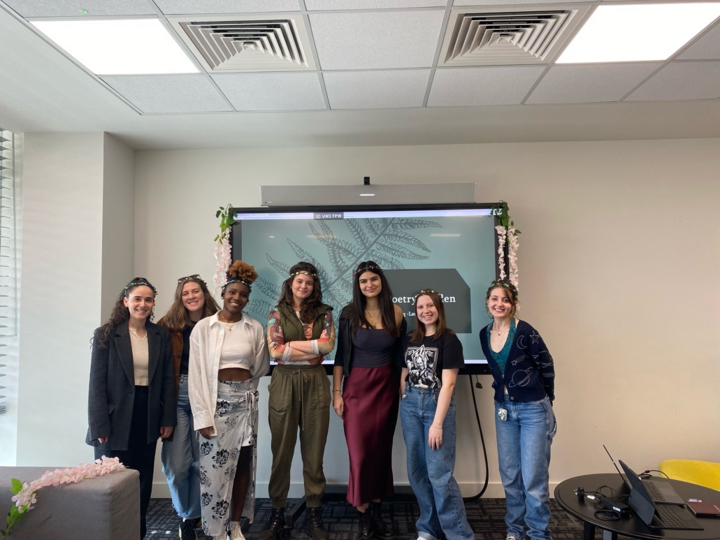 Seven people standing indoors, in front of a screen displaying "Poetry Garden." Floral decorations and casual attire create a relaxed atmosphere.