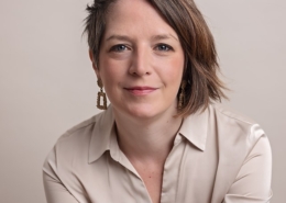 A person with short hair and earrings wears a beige shirt, posing against a neutral background, smiling gently at the camera.