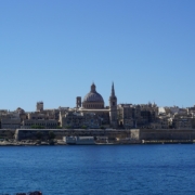 Skyline of Valletta, Malta with St. Paul's Cathedral and historic buildings in view against a clear blue sky, sea in foreground.