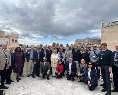 A group of people pose on a rooftop with the Acropolis in the background under a cloudy sky.