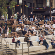 People relaxing on tiered seating by the river, enjoying a festival atmosphere with colorful bunting and food stalls nearby.