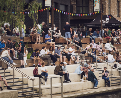 People relaxing on tiered seating by the river, enjoying a festival atmosphere with colorful bunting and food stalls nearby.