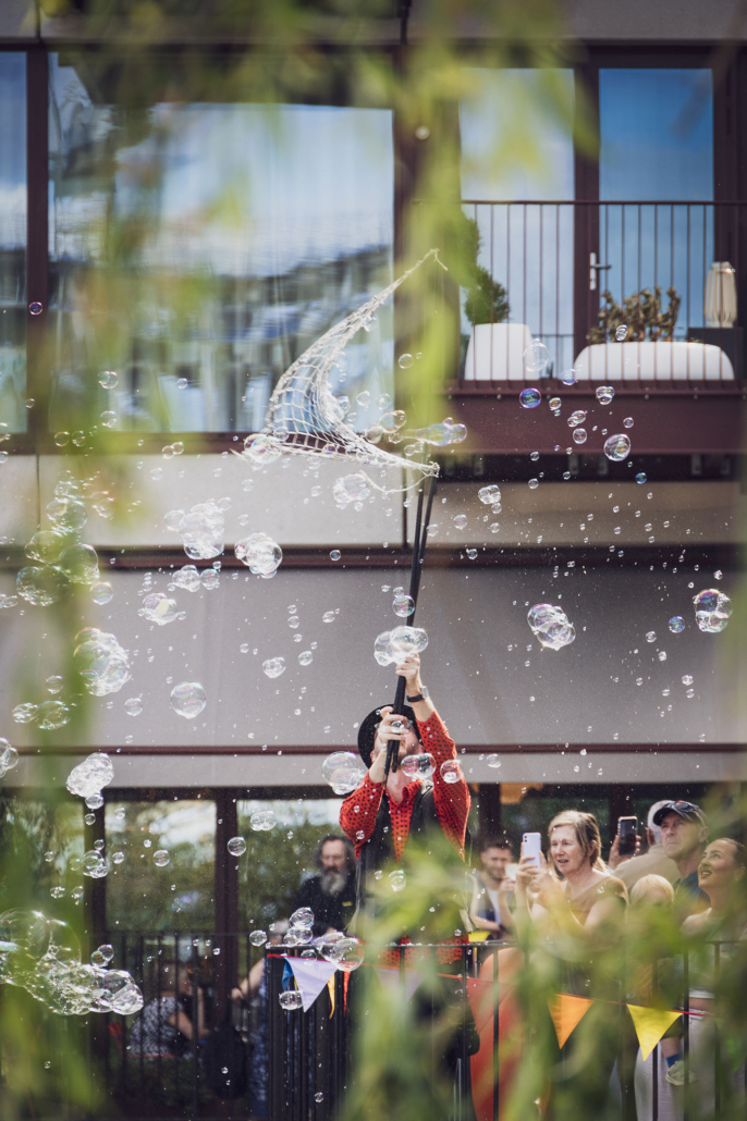 A person creates bubbles with a large net, surrounded by an audience capturing the moment with phones, in front of a building.