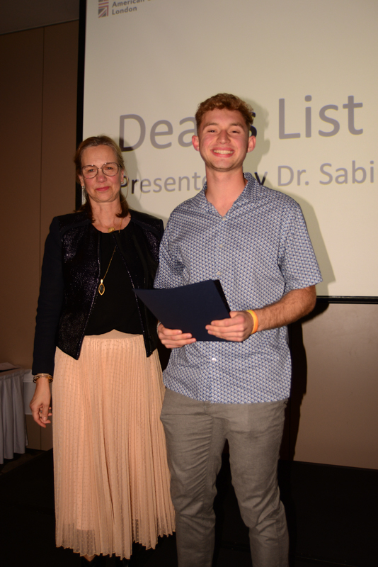 Two smiling people stand together near a presentation screen displaying "Dean's List" at an indoor event, one holding a certificate.