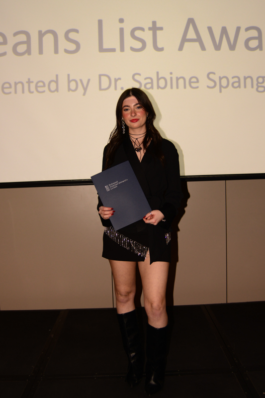 A person is standing on stage holding a certificate, with a presentation screen in the background displaying an award title.