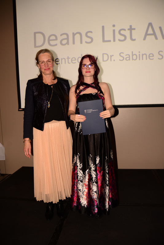Two people stand on a stage, one holding a certificate. Behind them, text reads "Deans List Award presented by Dr. Sabine S."
