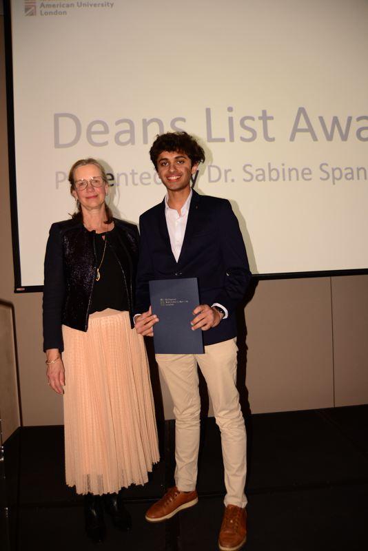 Two people stand on stage. One holds a certificate. "Dean's List Awards" is projected behind them at American University London.