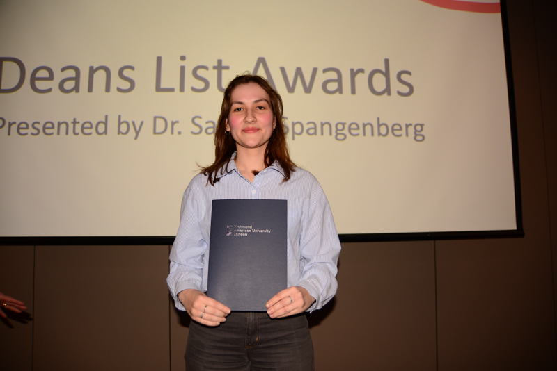 A person holding a certificate from Richmond American University London, standing in front of a "Dean's List Awards" presentation screen.