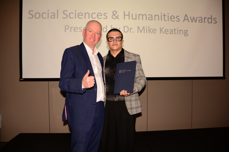 Two people stand smiling on a stage at an awards ceremony. One holds a certificate. A presentation screen is visible behind them.