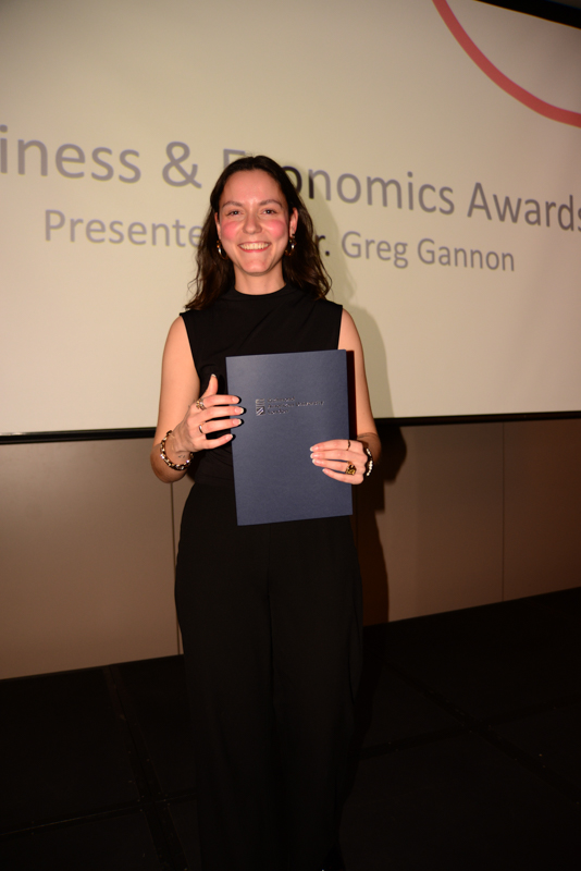 A person stands smiling on stage, holding an award certificate, with text indicating a Business & Economics Awards presentation in the background.