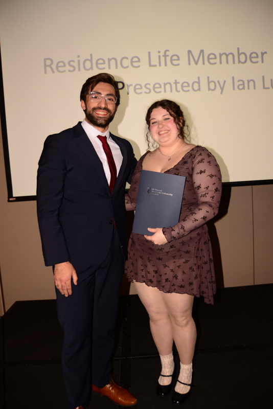 Two people stand on a stage, one holding a certificate. Behind them, a screen displays a presentation title related to Residence Life.