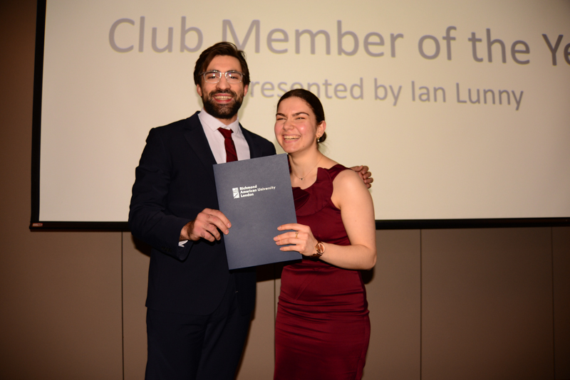 Two people smiling, holding a certificate at an award ceremony, with "Club Member of the Year" displayed on the screen behind them.