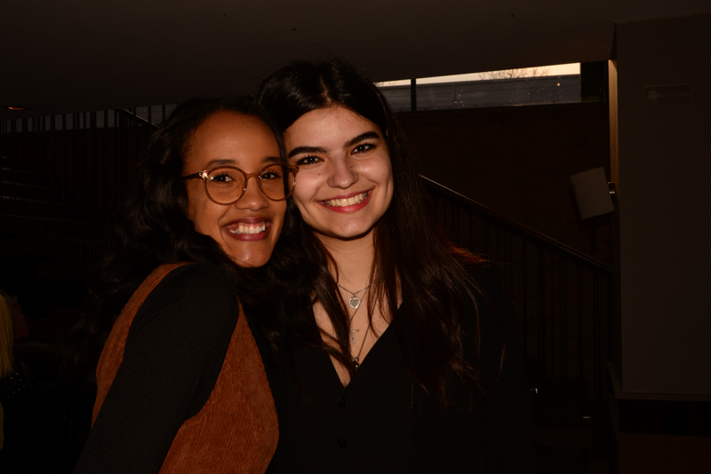 Two people smiling and posing indoors under dim lighting near a stairway, with a window in the background.