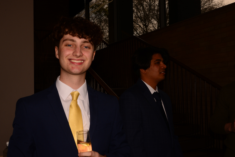 Two people in suits at an indoor gathering, one holding a drink and smiling, near a staircase with large windows behind them.