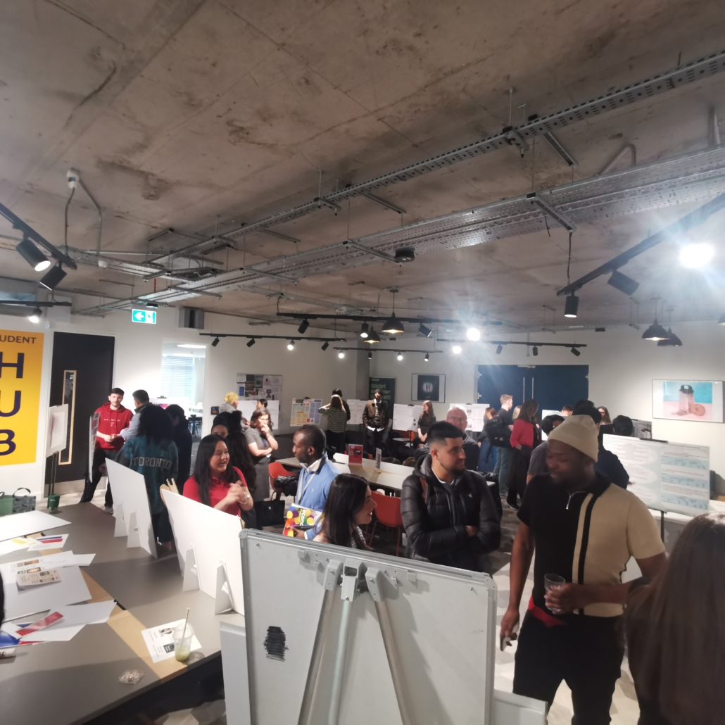 A group of people engage in discussions at an indoor exhibition or networking event, with posters and informational displays in the background.