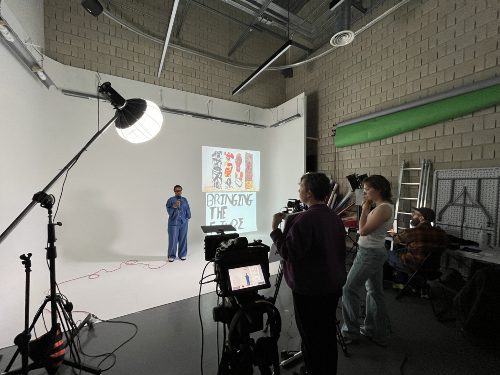 A person in blue clothing is speaking on a white set. Three people operate cameras and equipment, with a backdrop displaying artwork.