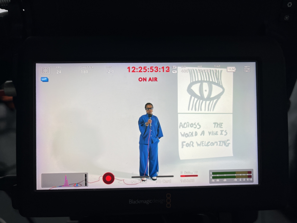 A person stands in front of a camera display, wearing blue, beside artwork and text reading, "Across the world, a vibe is for welcoming."