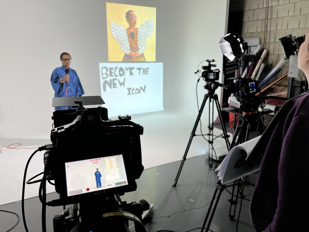 Person in blue shirt standing in a studio, holding microphone, with cameras and projection displaying "Become the New Icon."