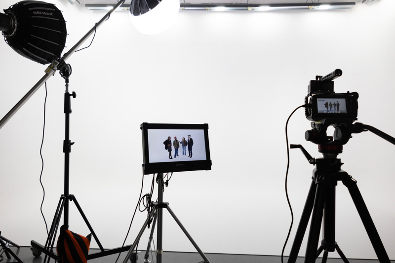 A photography studio setup with lights, camera, and monitor displaying four people on a white backdrop, ready for a photoshoot.