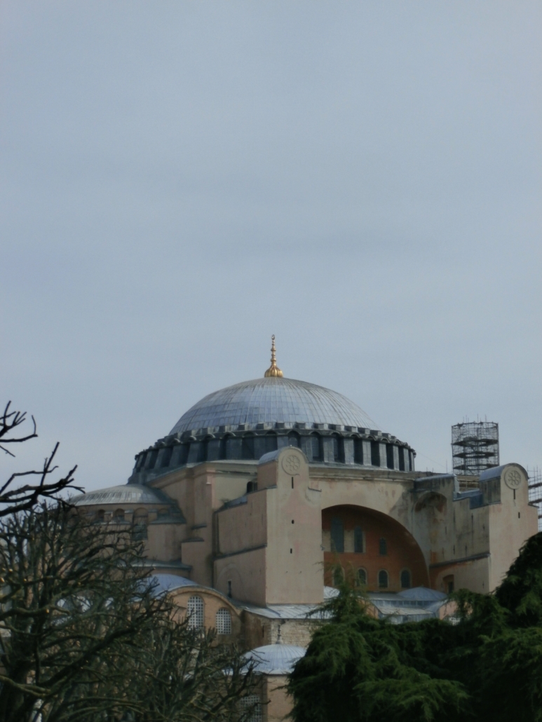 The image shows the Hagia Sophia’s grand dome, surrounded by trees and scaffolding against a cloudy sky, highlighting the historic architectural beauty.