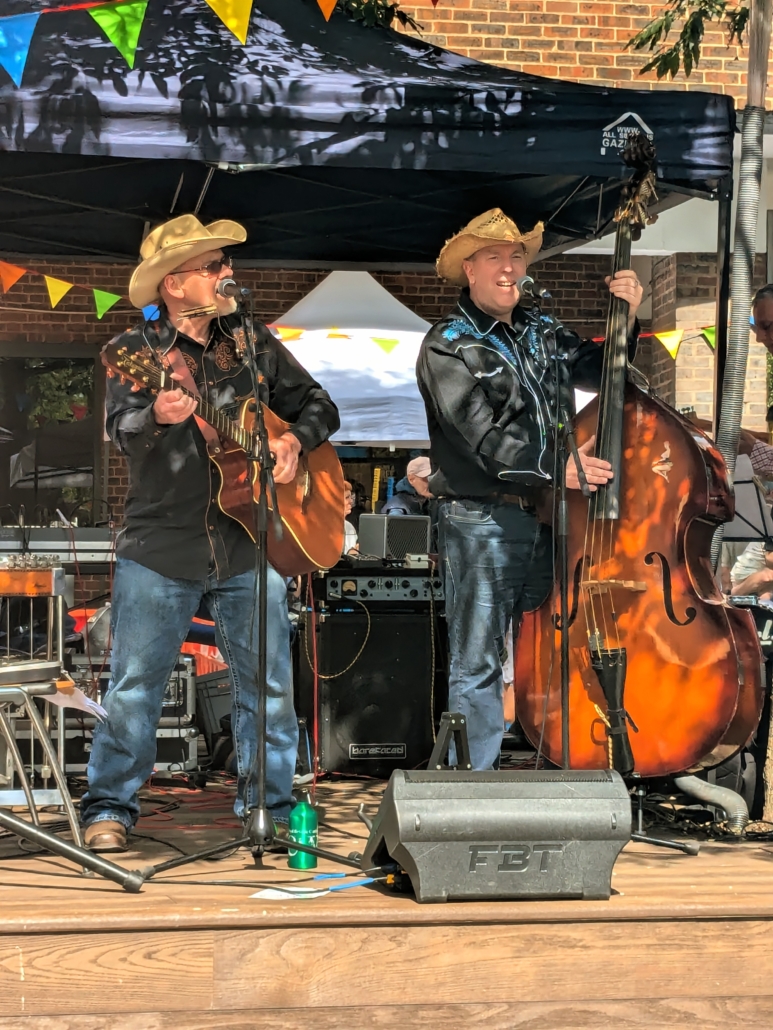 Two musicians in cowboy hats perform on stage with guitar and double bass under a canopy, surrounded by colorful flags and equipment.