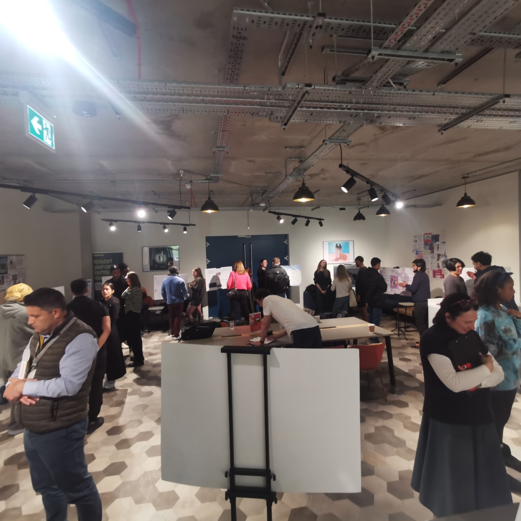 Several people are gathered in a modern exhibition space, engaging with displays and artwork. The room features industrial lighting and hexagonal tiles.