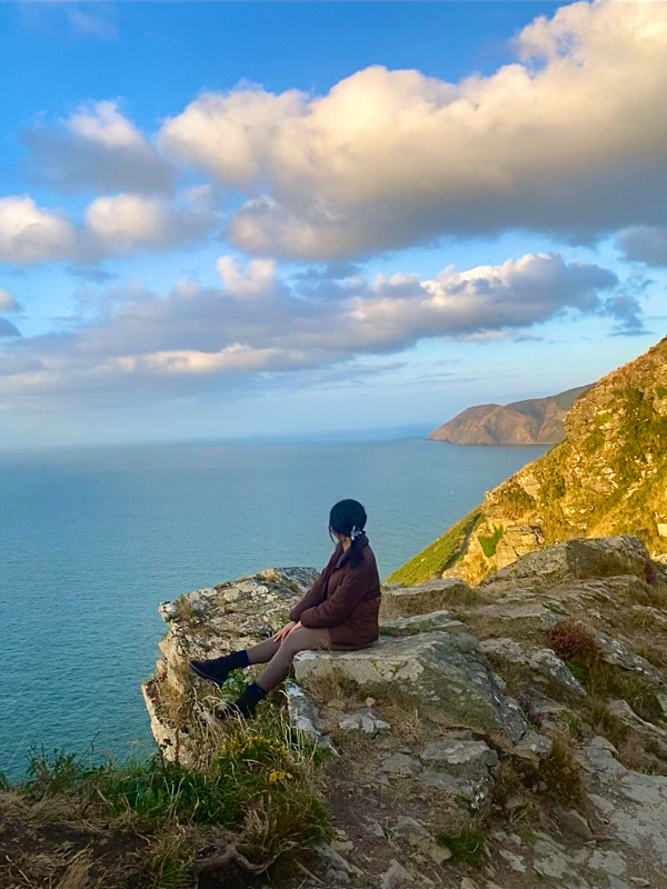 A person sits on a rocky cliff overlooking a vast ocean under a partly cloudy blue sky, surrounded by rugged coastal landscape.