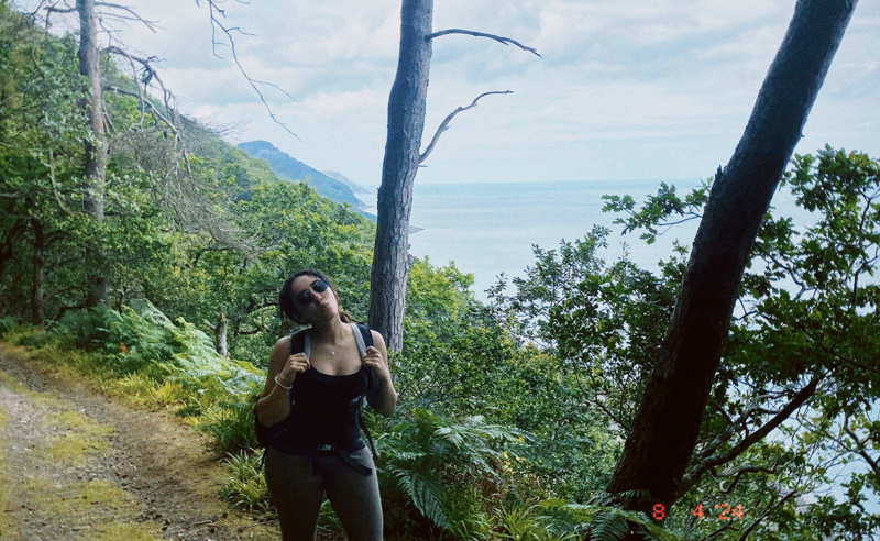 A person with a backpack poses on a forested path overlooking a scenic coastline, surrounded by lush greenery and distant rolling hills.