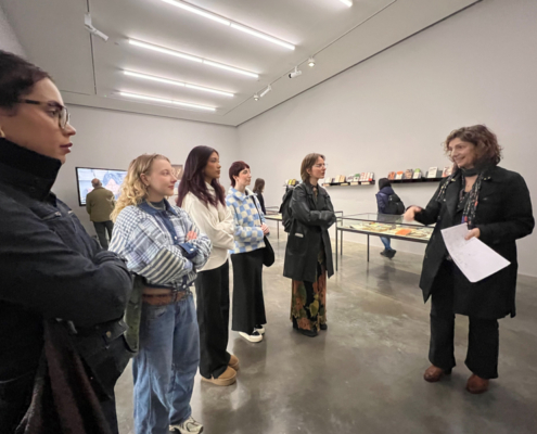 A group of people attentively listens to a presentation in a contemporary art gallery with paintings and documents on display.