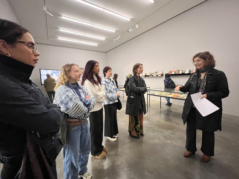 A group of people attentively listens to a presentation in a contemporary art gallery with paintings and documents on display.