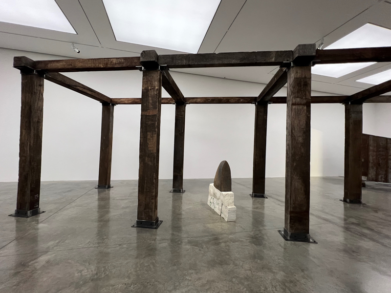 An art installation features wooden beams forming a square structure, with a stone and metal sculpture placed centrally on polished concrete flooring.