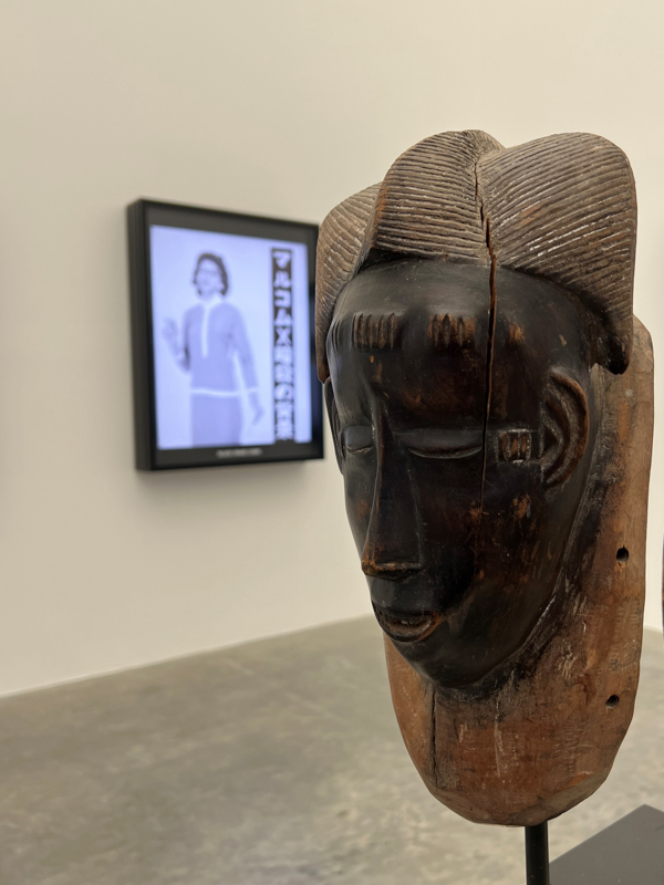 A wooden African mask is displayed in a gallery, with a blurred black-and-white image of a person in the background.