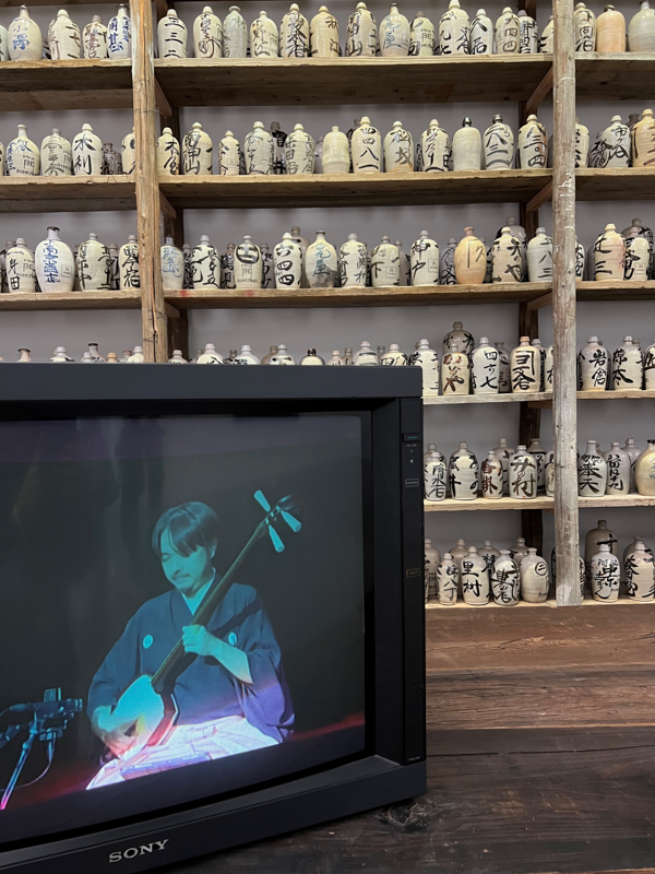 A person plays a string instrument on a television beside wooden shelves filled with labeled ceramic bottles, creating a vintage aesthetic.
