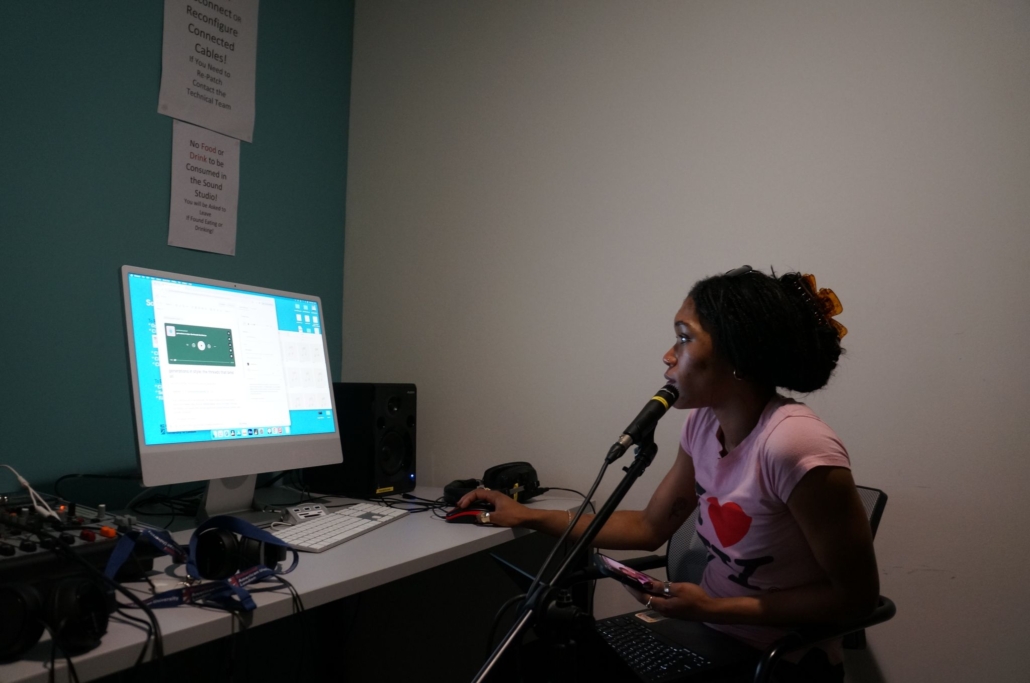 A person in a recording studio speaks into a microphone while using a computer. Cables and headphones are visible on the desk.