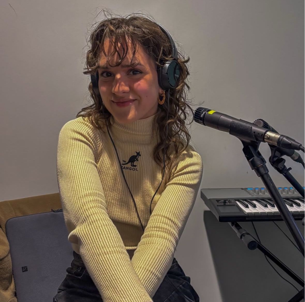 A person wearing headphones sits beside a microphone and keyboard. The person is smiling, wearing a cream sweater with a kangaroo logo.