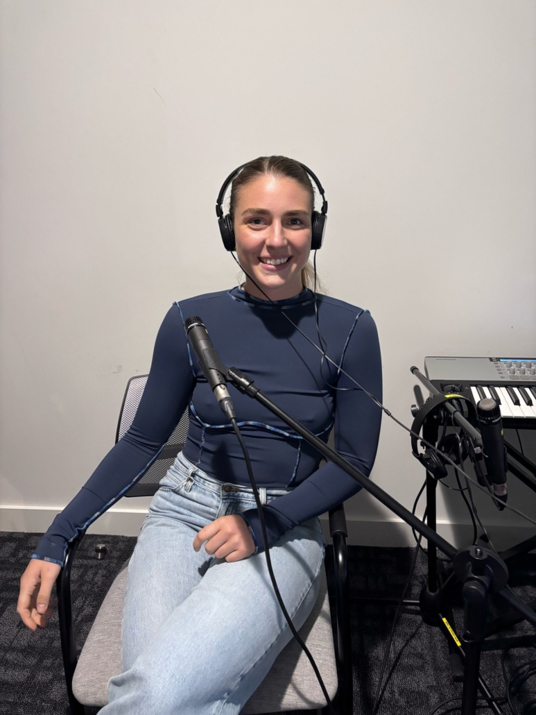 Person in a blue shirt and headphones, seated with a microphone, near a keyboard. Plain white wall background.