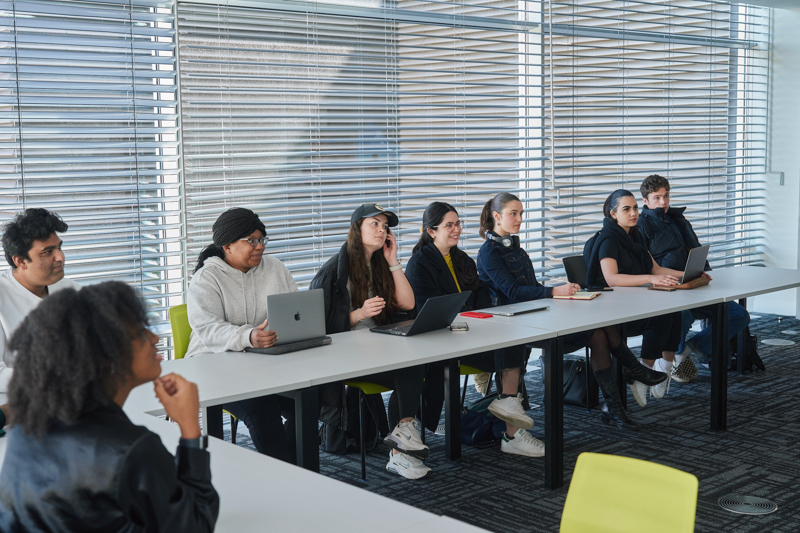 A group of people seated in a modern classroom, using laptops, engaged in a discussion. Blinds cover the large windows behind them.