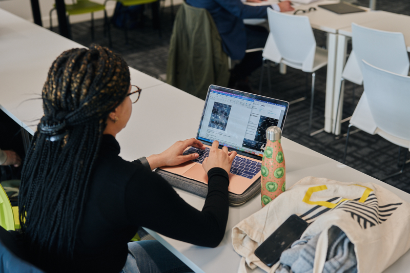A person works on a laptop in a classroom setting, with a reusable bottle and bag nearby, surrounded by white chairs and desks.
