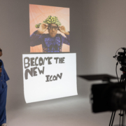 A person in a studio holds a microphone, facing a poster that reads "Become the New Icon" with a colorful image above.