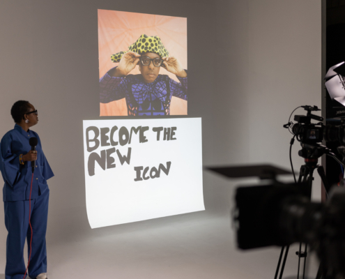 A person in a studio holds a microphone, facing a poster that reads "Become the New Icon" with a colorful image above.