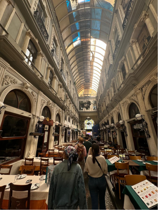 An ornate indoor arcade with high glass ceiling, featuring dining tables, elaborate architecture, and a few people walking among the restaurant seating.