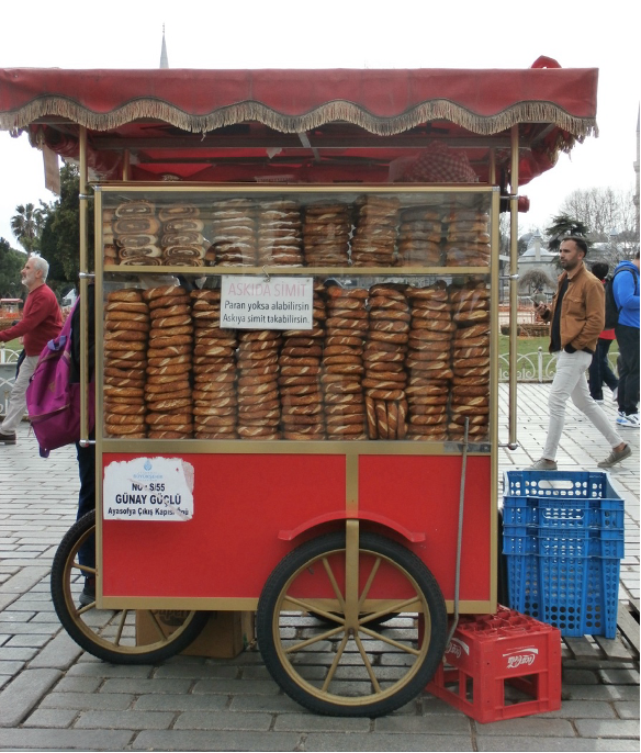 A red simit cart filled with bagels stands on a cobblestone path by Hagia Sophia, with two people walking nearby.