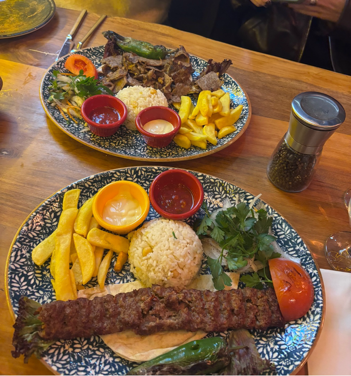 Two plates on a wooden table with kebabs, rice, fries, greens, and dips. A pepper grinder is seen nearby.