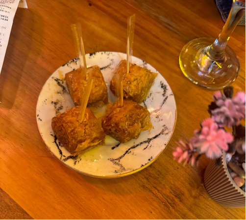 A plate of four small glazed pastries on a wooden table, with skewers inserted. Nearby, a wine glass and a small flower pot.