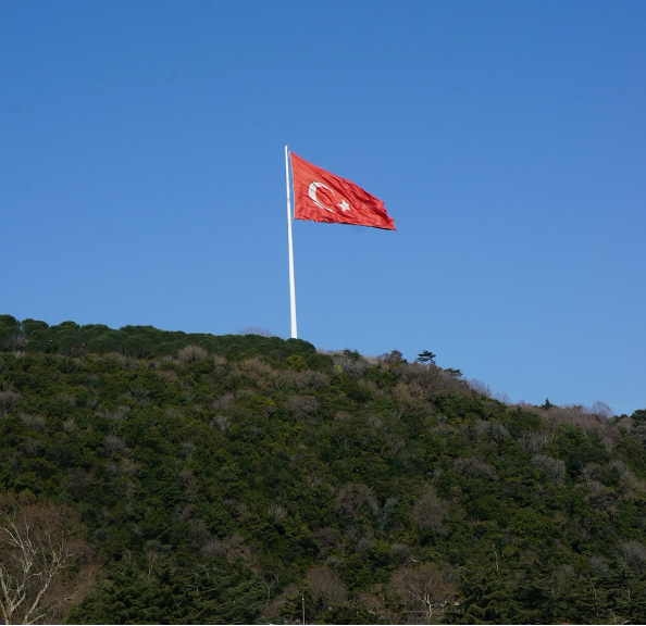 A large Turkish flag on a hilltop flutters against a clear blue sky, surrounded by dense, green foliage.