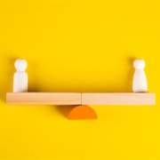 Two wooden figures balance on a seesaw-like wooden structure, against a bright yellow background, symbolizing equilibrium or comparison.