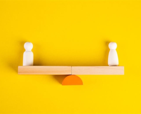 Two wooden figures balance on a seesaw-like wooden structure, against a bright yellow background, symbolizing equilibrium or comparison.