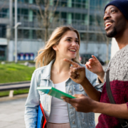 Two people smile and converse outdoors, holding maps. They're in a modern urban setting with greenery and buildings in the background.