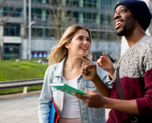 Two people smile and converse outdoors, holding maps. They're in a modern urban setting with greenery and buildings in the background.