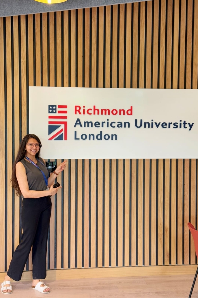A person is pointing at a sign for Richmond American University London, standing against a wood-paneled wall.